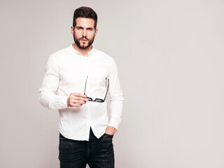 Portrait of handsome confident  model. Sexy stylish man dressed in white shirt and jeans. Fashion hipster male posing in studio on grey background. Taking off sunglasses. Isolated