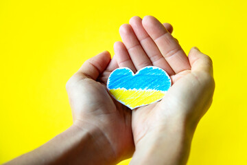 woman's hands holding a paper heart cut out in the colors of the Ukrainian flag on yellow background with space for copy. War.