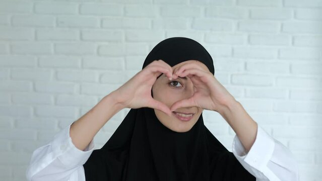 A Muslim woman in a black hijab smiles sweetly and shows a sign of love in the form of a heart folded with her fingers. A symbol of the Islamic world's love for the whole world.
