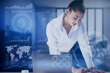 Attractive young european female using laptop at desktop with abstract global digital business interface on blurry background. Accounting, chart, finance and hud concept. Double exposure.