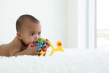 Adorable African Asian newborn girl lying on bed holding and biting toy at home. Baby teething...