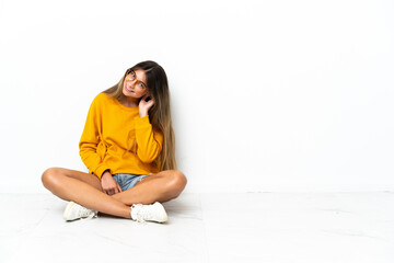Young woman sitting on the floor isolated on white background listening to something by putting hand on the ear