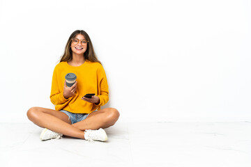 Young woman sitting on the floor isolated on white background holding coffee to take away and a...