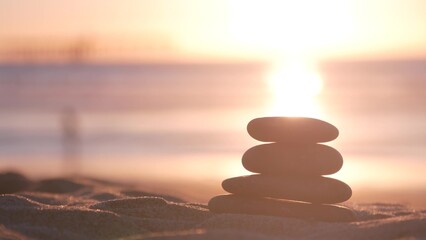 Stack of pebble stones, sandy ocean beach, sunset sky. Rock balancing in sun light, sea water waves. Stones staking in pyramid pile. Zen meditation and harmony in balance. Calm tranquil atmosphere.