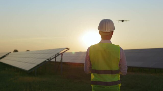 Back view of man in helmet and safety vest activating drone for survey and exploration solar panels on station. Modern technology for work. Production of alternative energy.