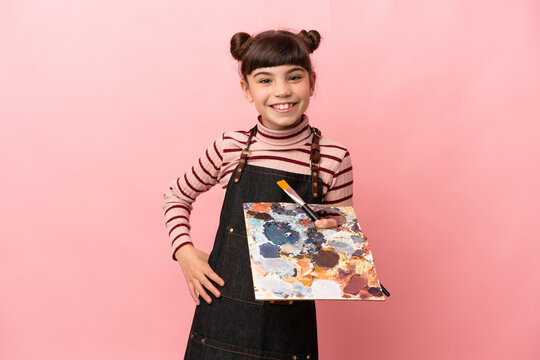 Little artist girl holding a palette isolated on pink background posing with arms at hip and smiling