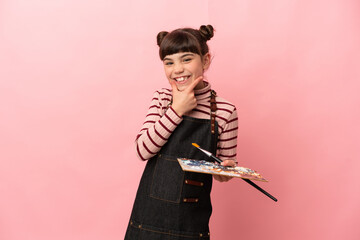 Little artist girl holding a palette isolated on pink background happy and smiling