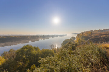 Autumn landscape in the early morning overlooking the river. A wide river and endless expanses of fields. Yellow leaves on trees and bushes are illuminated by the rays of the rising sun.