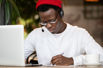 Focused African American young guy freelancer analyst in wireless headphones works with documents...