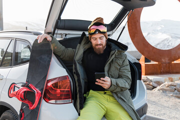 Young man with snowboard sitting in car trunk using smart phone