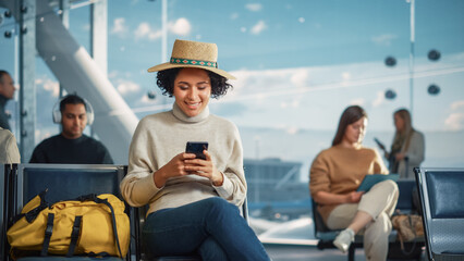 Airport Terminal: Happy Black Woman Waits for Flight, Uses Smartphone, Browse Internet, Post on...