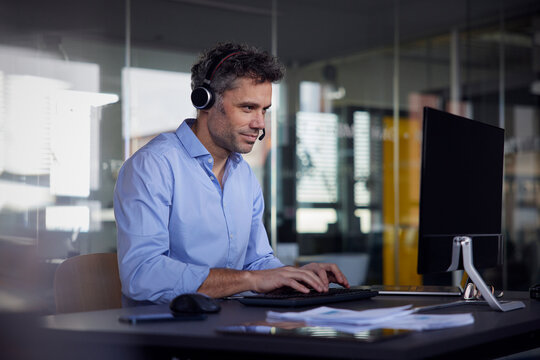 Businessman wearing wireless headset using computer sitting at desk in office