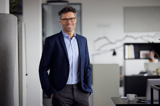 Happy Businessman With Hands In Pockets Standing By Desk In Office