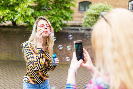Teenager photographing friend blowing soap bubbles through smart phone