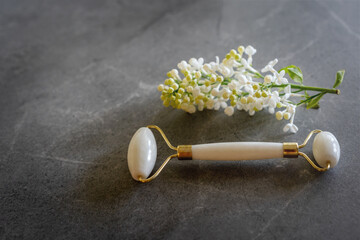 Jade facial roller – skincare lifting and toning tool. White beauty roller, massager and lily of the valley flowers on a grey background.