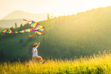 Little boy in hat at sunset runs colored striped kite. Wonderful mountain landscape with ridges and fir trees. Sunny summer or spring day. Summer vacation and healthy lifestyle concept. Copy space.