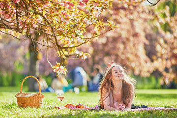 Beautiful young woman having picnic on sunny spring day in park during cherry blossom season