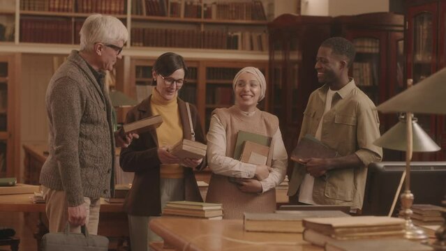 Medium slowmo portrait of cheerful diverse group of people with books in hands having conversation standing at public library with wooden furniture then smiling at camera