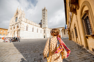 Woman enjoys beautiful architecture of Siena cathedral, standing back on Duomo square in Siena town. Traveling old towns of Tuscany region of Italy. Woman wearing colorful shawl in hair