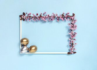 Easter eggs in silver and gold color with pink flowers. Creative concept in white card frame on blue background. Minimal flat lay.