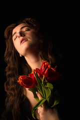 Studio portrait of a young woman in sunglasses, on a black background, with a bouquet of red tulips in her hands. International Women's Day. Valentine's Day