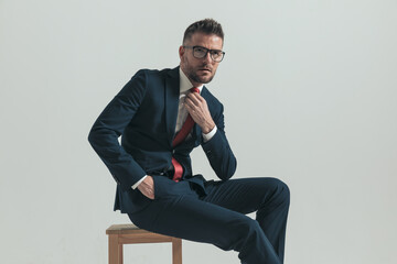 confident unshaved man in elegant suit fixing tie and looking up