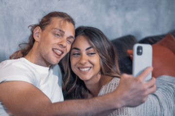 Cheerful couple of romantic caucasian guys make face taking a selfie at night in living room messaging and watching their cellphone device. Male fun guy enjoyng life looking a smart phone with a girl