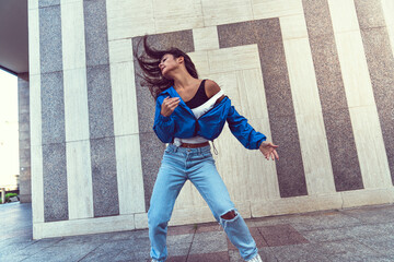 Photo shoot of stylish caucasian hip hop dancer posing having fun in blue jacket, jeans, black top and white tshirt, enjoy the urban town as background. City buildings shooting of moving fitness model