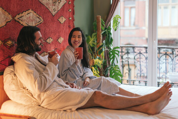 Obraz premium multiethnic couple in bathrobe lying on the bed enjoying a cup of coffee or tea or herbal tea in the morning
