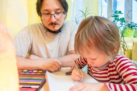 Bearded man in grey t-shirt and cute boy drawing pictures with colored wax crayons while spending time at home together.