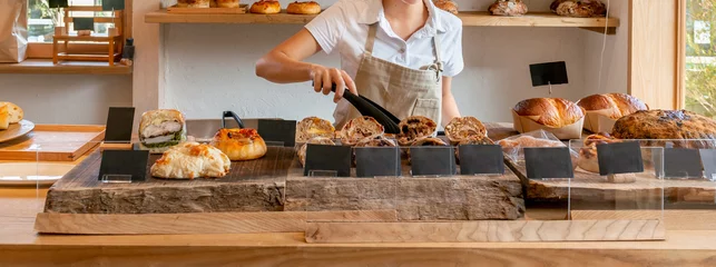 Foto op Plexiglas パン屋　ベーカリーショップ　トングで焼きたてパンを掴むエプロン姿の女性店員 © Metro Hopper