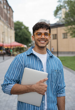 Authentic portrait of young handsome Indian man wearing stylish eyeglasses standing on the street. Smiling asian student holding laptop looking at camera in university campus. Education concept 