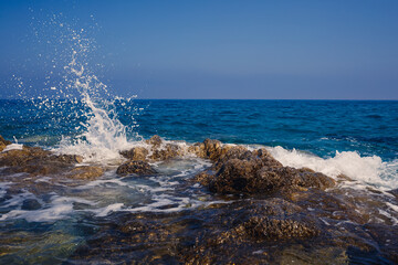 Wonderful views of the blue Mediterranean Sea. Sunny rocks, waves with foam and splashing water. The wave crashes into the rocks on the shore