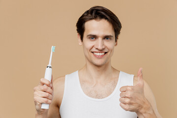 Attractive young man 20s perfect skin wearing undershirt hold brush brushing teeth show thumb up...