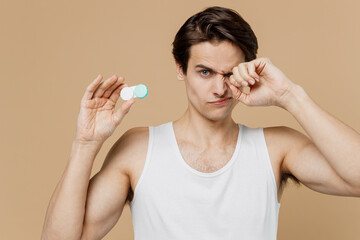 Attractive sad young man 20s perfect skin in undershirt hold contact lenses case point finger on container rub his eye isolated on pastel pastel beige background Eyecare healthcare procedures concept