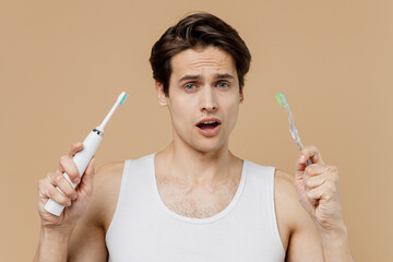 Attractive puzzled confused young man 20s perfect skin in undershirt hold choice electric brushes...