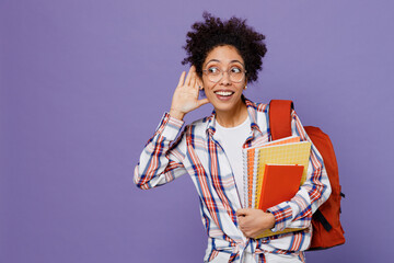 Fototapeta premium Young girl woman of African American ethnicity student in shirt backpack try hear you overhear listen intently isolated on plain purple background. Education in high school university college concept.