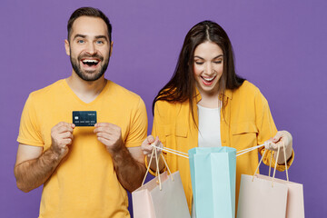Young excited happy couple two friends family man woman together in yellow casual clothes hold package bags with purchases after shopping credit bank card isolated on plain violet background studio.