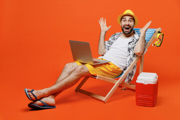 Young surprised fun tourist man wear beach shirt hat lie on deckchair hold use work on laptop pc computer isolated on plain orange background studio portrait. Summer vacation sea rest sun tan concept