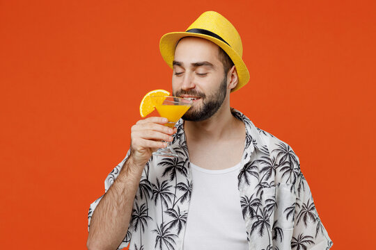 Young tourist man wear beach shirt hat travel abroad on weekends hold sniff alcohol cocktail close eyes isolated on plain orange background studio portrait Summer vacation sea rest sun tan concept