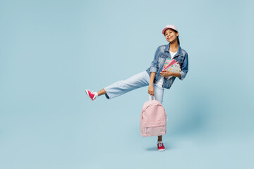 Full body fun little schoolgirl kid teen girl of African American ethnicity 12-13 year old in denim jacket backpack cap hold exercise books raise up leg isolated on pastel plain light blue background.