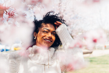 Happy and carefree Brazilian woman with afro hair in spring with pink flowers around her. Woman...