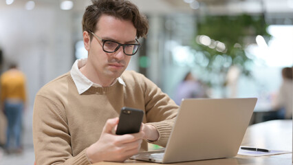 Young Man using Smartphone while Working on Laptop