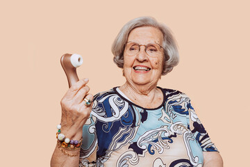 Funny happy smiling elderly grandmother holding satisfyer dildo with hand at studio over beige...