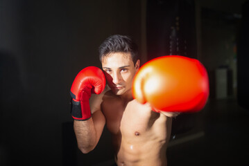 Portrait of handsome young boxer kicking at camera. Shirtless man in red boxing gloves training in dark space. Strength concept