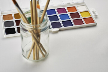 watercolor paints and a jar with brushes for painting on a white surface
