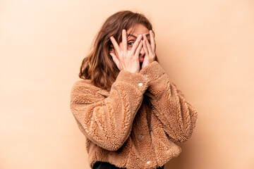 Young caucasian woman isolated on beige background blink through fingers frightened and nervous.