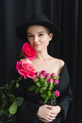 Fototapeta premium young woman in black blazer and brim hat looking at camera near fresh roses on dark background.
