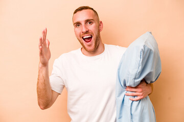 Young caucasian man wearing a pajama holding pillow isolated on beige background receiving a...