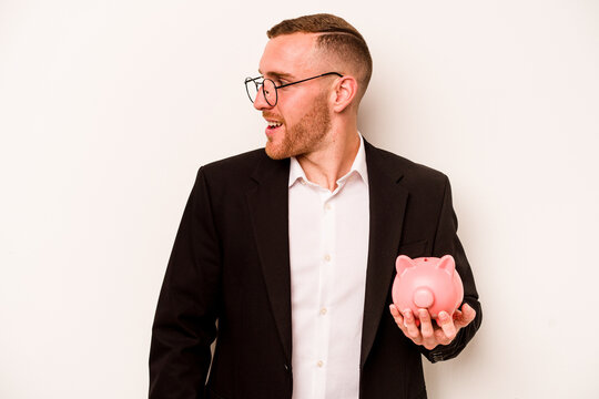 Young business caucasian man holding piggy bank isolated on white background looks aside smiling, cheerful and pleasant.